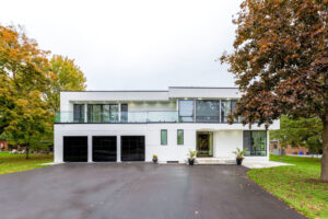 A modern two-story white house with large windows, a glass balcony, and a paved driveway, surrounded by green grass and trees.