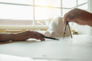Two people review architectural blueprints on a table, with one holding a compass and a hard hat and rolled-up plans in the background.