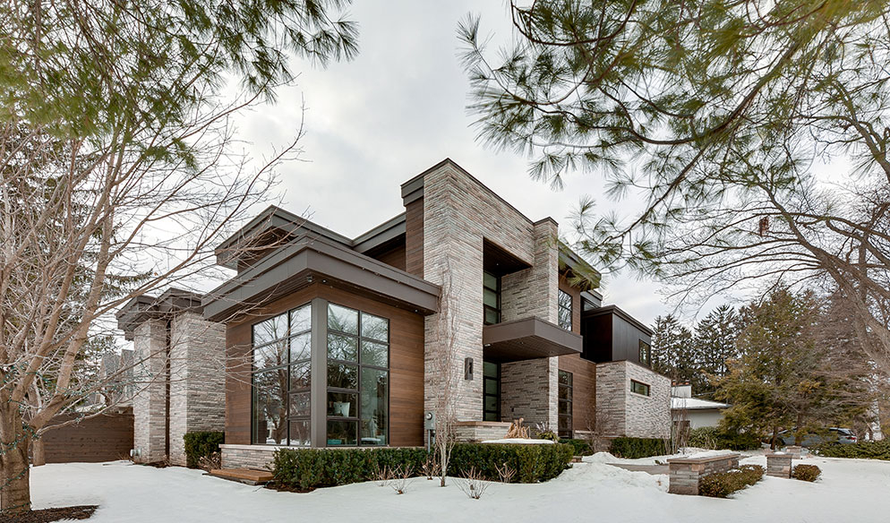 Modern two-story house with large windows, stone and wood exterior, surrounded by snow-covered ground and trees on a cloudy day.