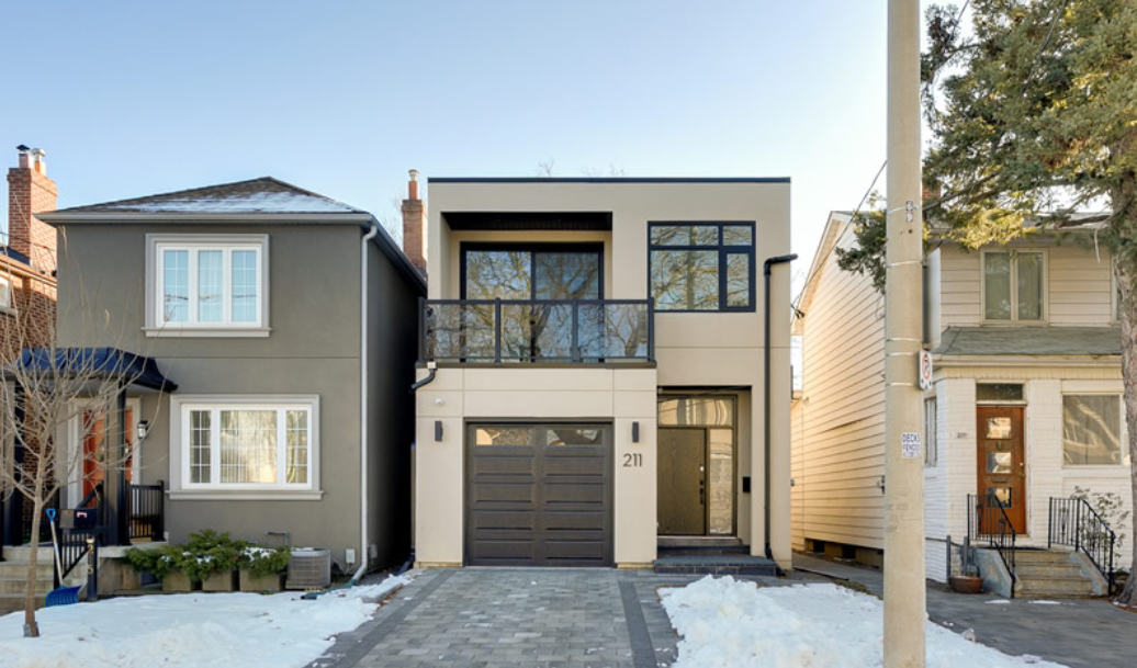Modern two-story house with flat roof, large windows, and attached garage, situated between two older homes on a winter day with snow on the ground. House number 211 is visible.