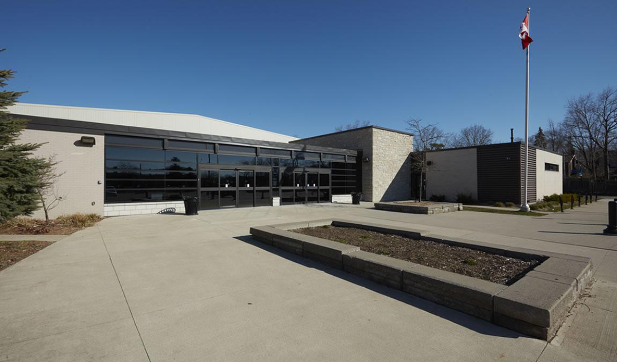 A modern, single-story building with large windows, a concrete courtyard, empty garden beds, and a Canadian flag on a flagpole under a clear blue sky.