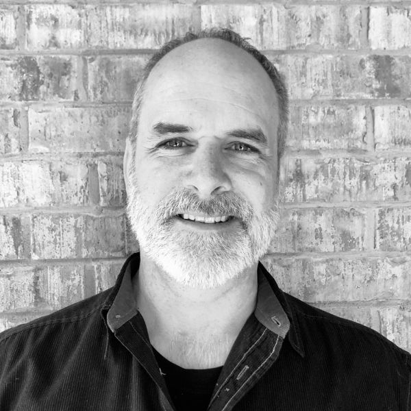 A middle-aged man with a short beard and receding hairline smiles in front of a brick wall. He is wearing a dark, collared shirt.