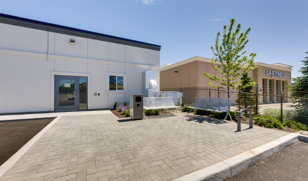 A paved walkway with benches and a small tree outside a white building, with an LA Fitness gym visible in the background.