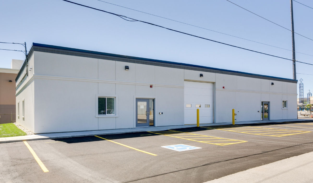 Single-story industrial building with white walls, several doors and windows, a large garage door, and a parking lot with marked spaces, including one accessible parking spot.