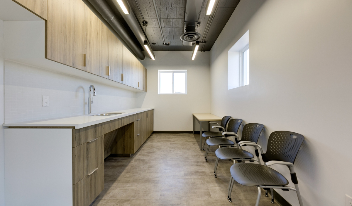 A modern break room with wood cabinets, sink, countertop, and four black chairs lined up along the opposite wall under a window.