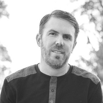 A man with short hair and a beard, wearing a buttoned long-sleeve shirt, stands outdoors with blurred trees in the background.