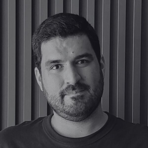 Man with short dark hair and beard facing forward, wearing a dark shirt, standing in front of vertical striped background. Black and white photo.