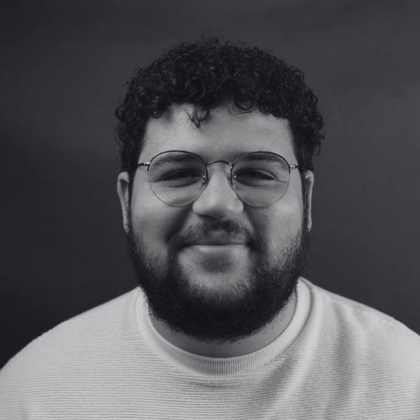 A person with curly hair, glasses, and a beard is smiling at the camera against a plain dark background.