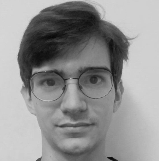 A young person with short dark hair and round glasses looks directly at the camera against a plain background.