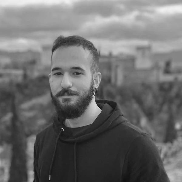 A bearded person with short hair and an ear piercing stands outdoors wearing a dark hoodie, with blurred buildings and trees in the background. The photo is in black and white.