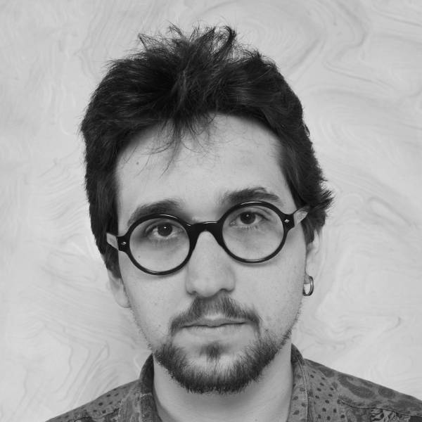 A person with short dark hair, round glasses, a mustache, and a beard is looking at the camera, wearing an earring and patterned shirt in front of a light marbled background.