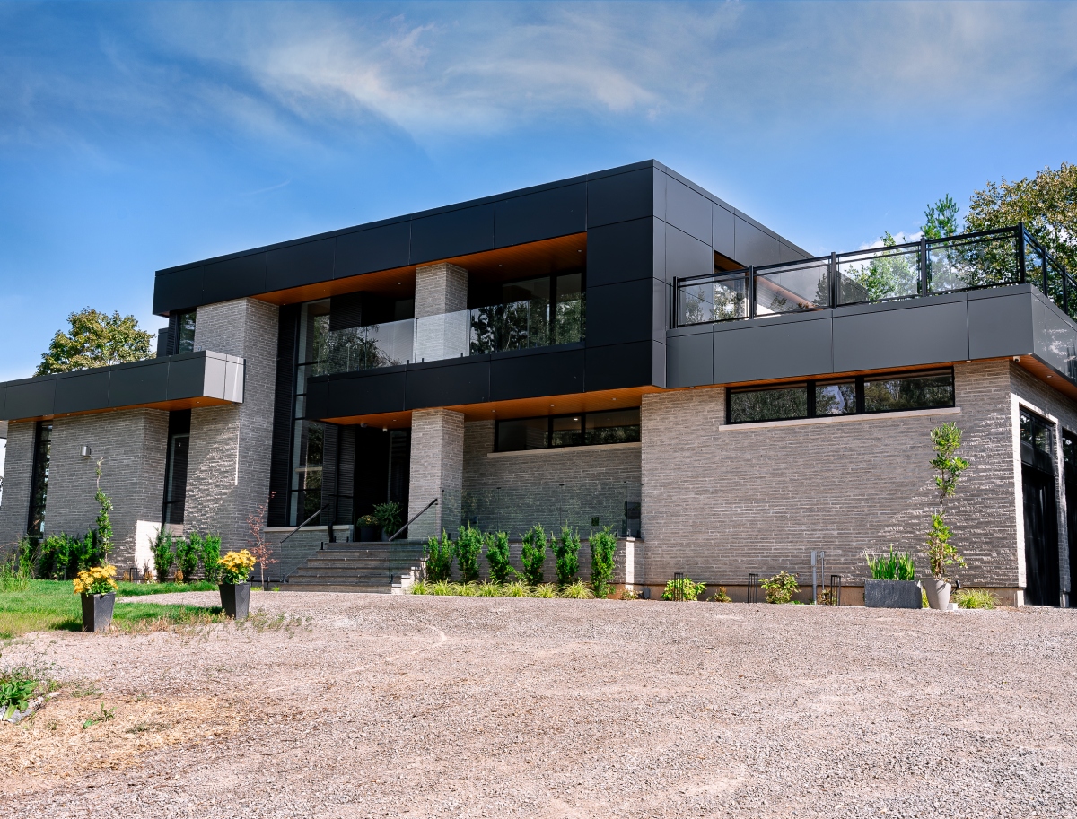 A modern two-story house with large windows, gray brick and black panel exterior, and a spacious front yard with plants and gravel.
