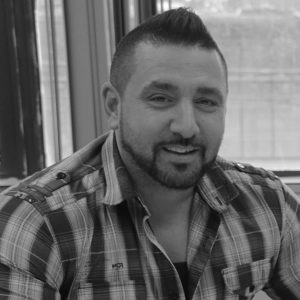 Man with short dark hair and a beard, wearing a plaid shirt, sitting indoors near a window, smiling at the camera.
