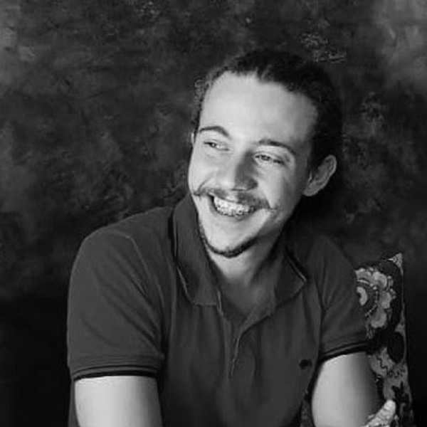 A young man with a beard and mustache smiles while sitting, wearing a short-sleeve polo shirt against a dark, textured background.