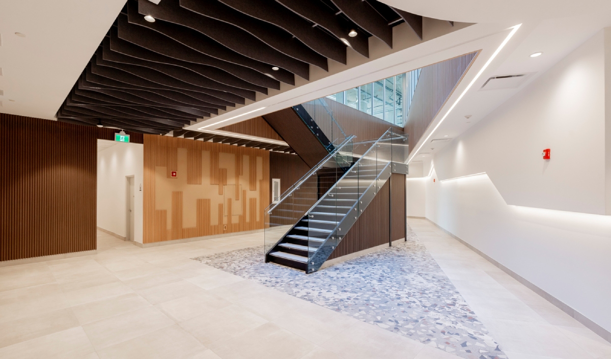 Modern building interior with tiled floors, wood panel walls, and a glass railing staircase leading to the upper floor under a black geometric ceiling.