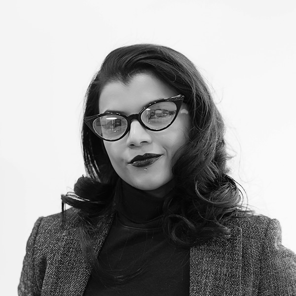 Black-and-white portrait of a woman with wavy hair, wearing glasses, a dark turtleneck, and a textured blazer, looking at the camera with a neutral expression.