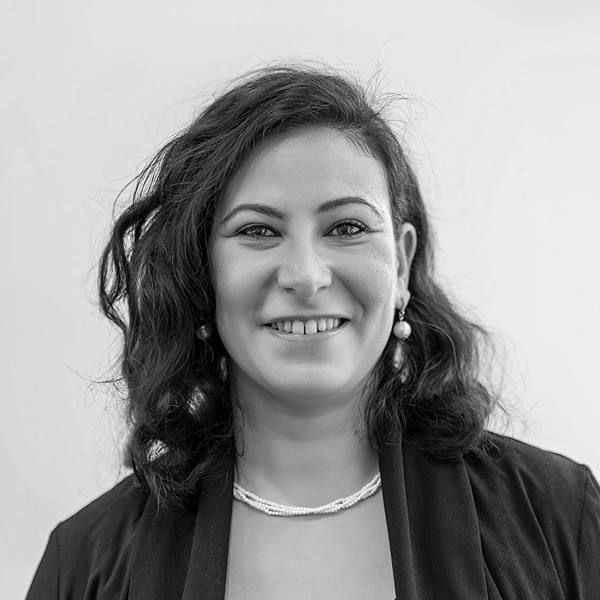 A woman with wavy dark hair, wearing a dark blazer, a necklace, and earrings, smiles at the camera against a plain light background.