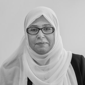 A woman wearing glasses and a white hijab, dressed in a dark blazer, faces the camera against a plain background.