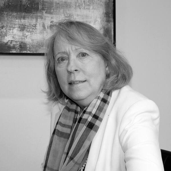 A woman with shoulder-length hair wearing a white blazer and a plaid scarf sits indoors in front of an abstract painting.