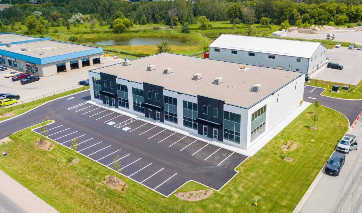 Aerial view of a modern commercial building with a mostly empty parking lot, surrounded by grass and neighboring industrial structures.