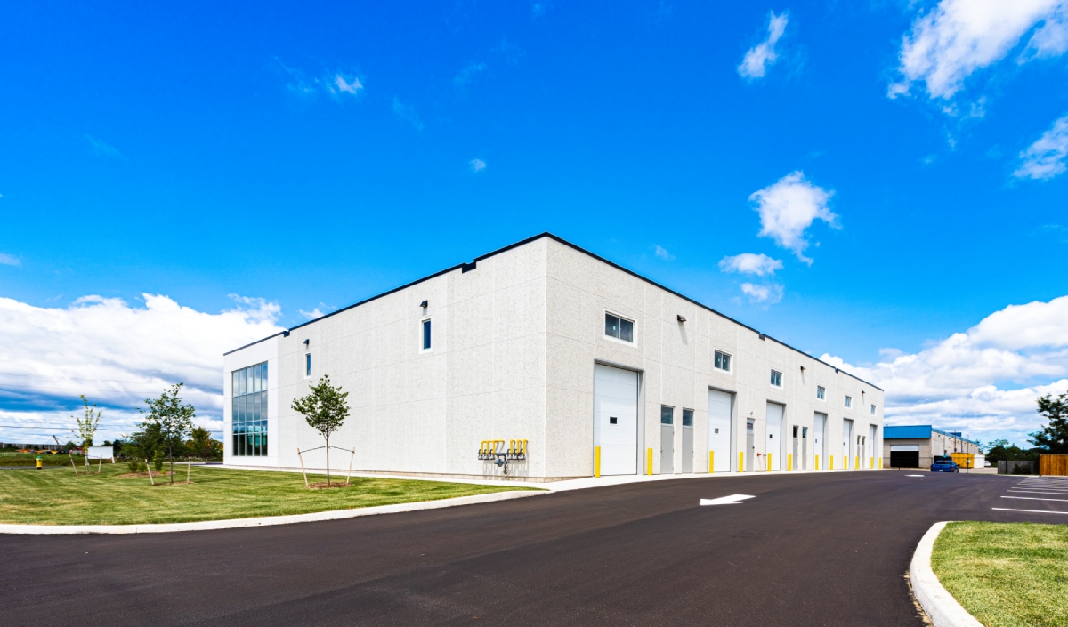 Large white industrial warehouse with multiple garage doors, surrounded by grass and a paved driveway, under a partly cloudy blue sky.