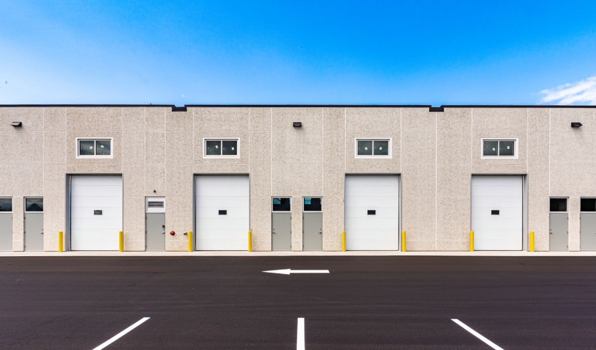 A row of white industrial garage doors on a beige warehouse building with an empty asphalt parking lot and a white arrow painted on the ground.