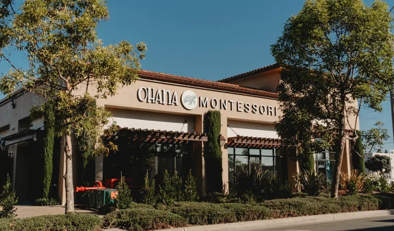 A single-story building with a sign reading “Ohana Montessori,” surrounded by trees and shrubbery under a clear blue sky.