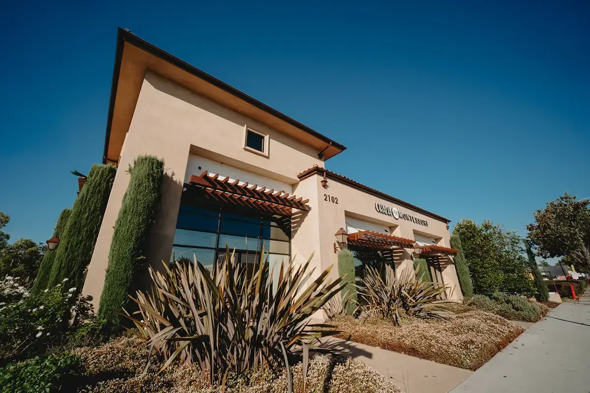 A beige stucco building with large windows, brown wooden awnings, and landscaping in front, under a clear blue sky.