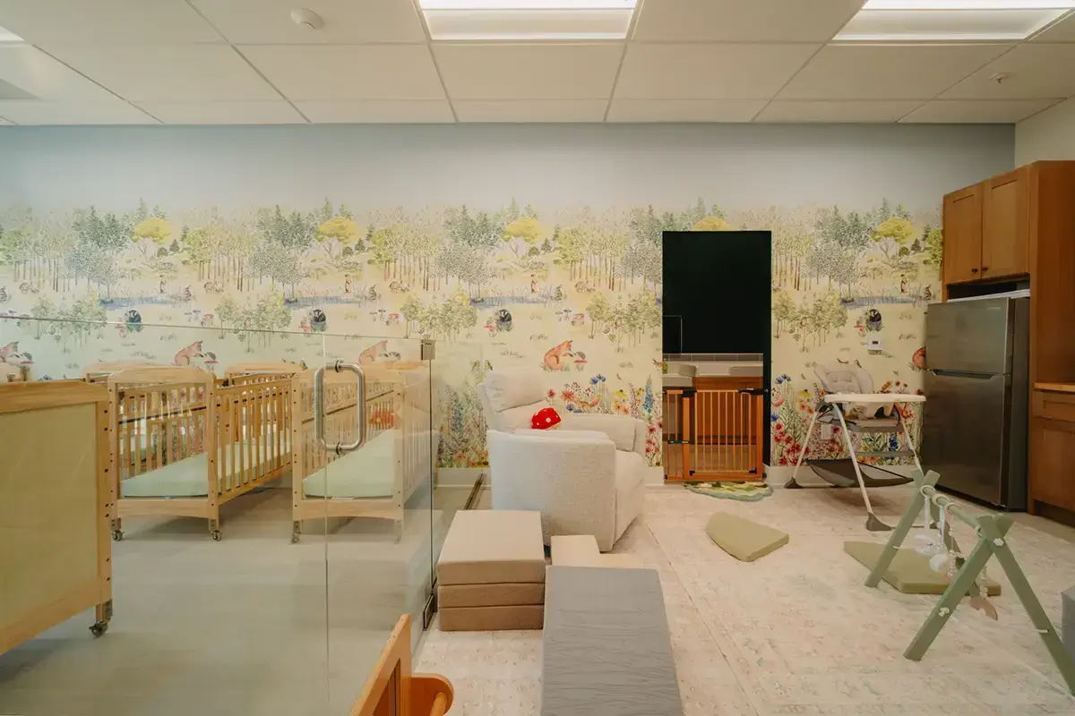A nursery room with cribs, a white armchair, play mats, a high chair, a fridge, cabinets, and a wallpaper featuring a nature scene. The room is well-lit and organized.