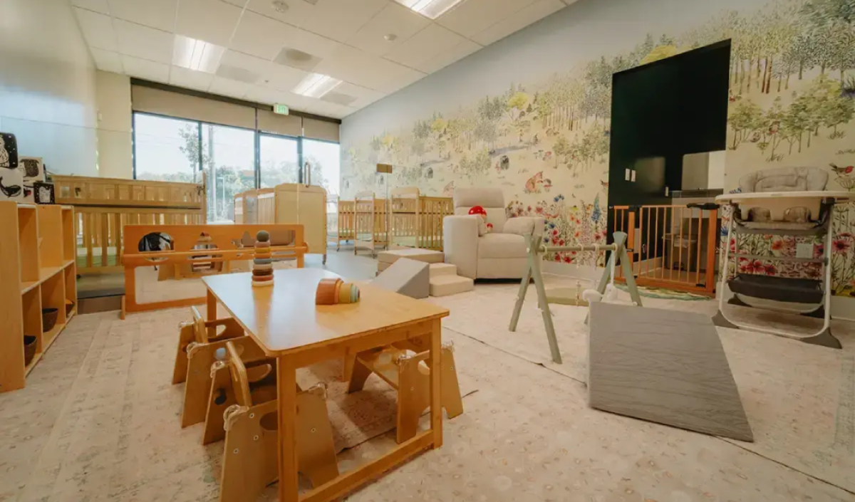 A daycare room with wooden tables, small chairs, cribs, toys, play mats, and a mural of trees on the back wall. Large windows let in natural light.