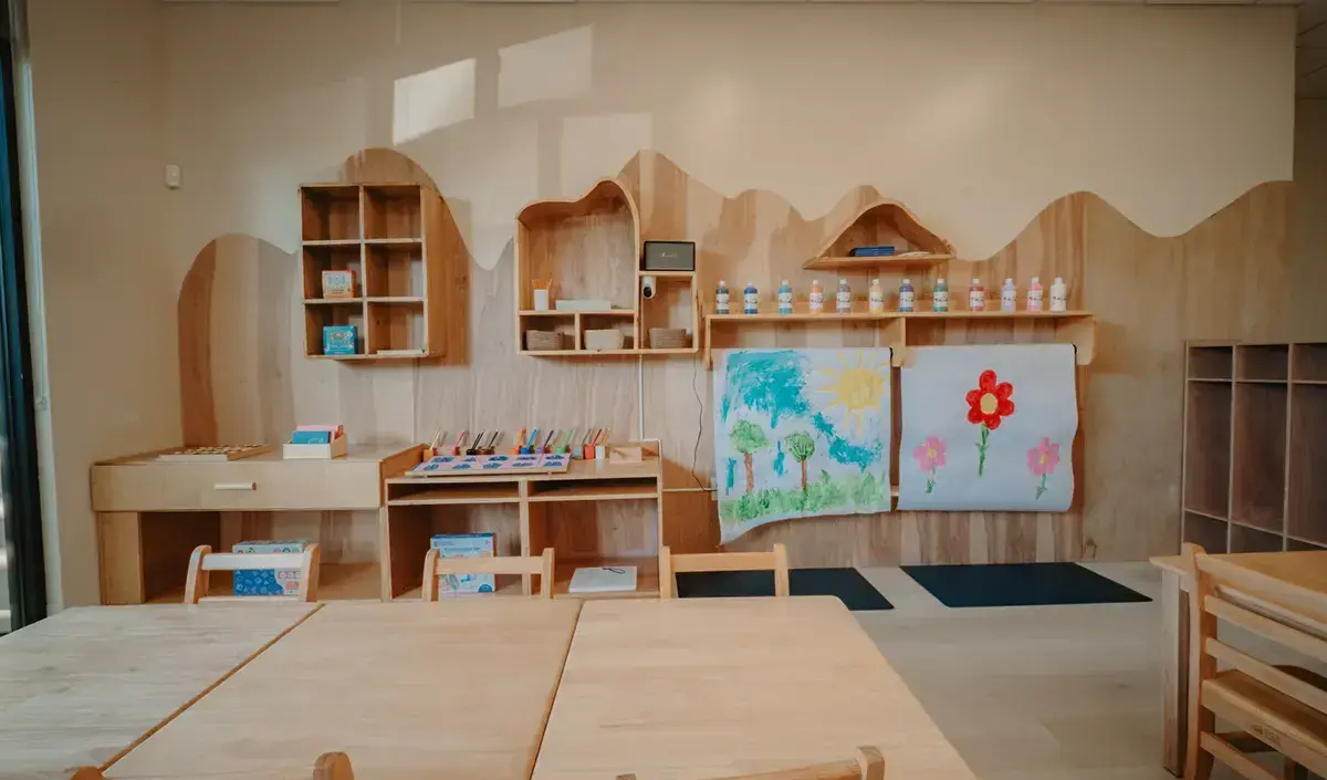 A classroom with wooden shelves, tables, art supplies, children’s paintings on the wall, and bottles arranged neatly on a shelf.
