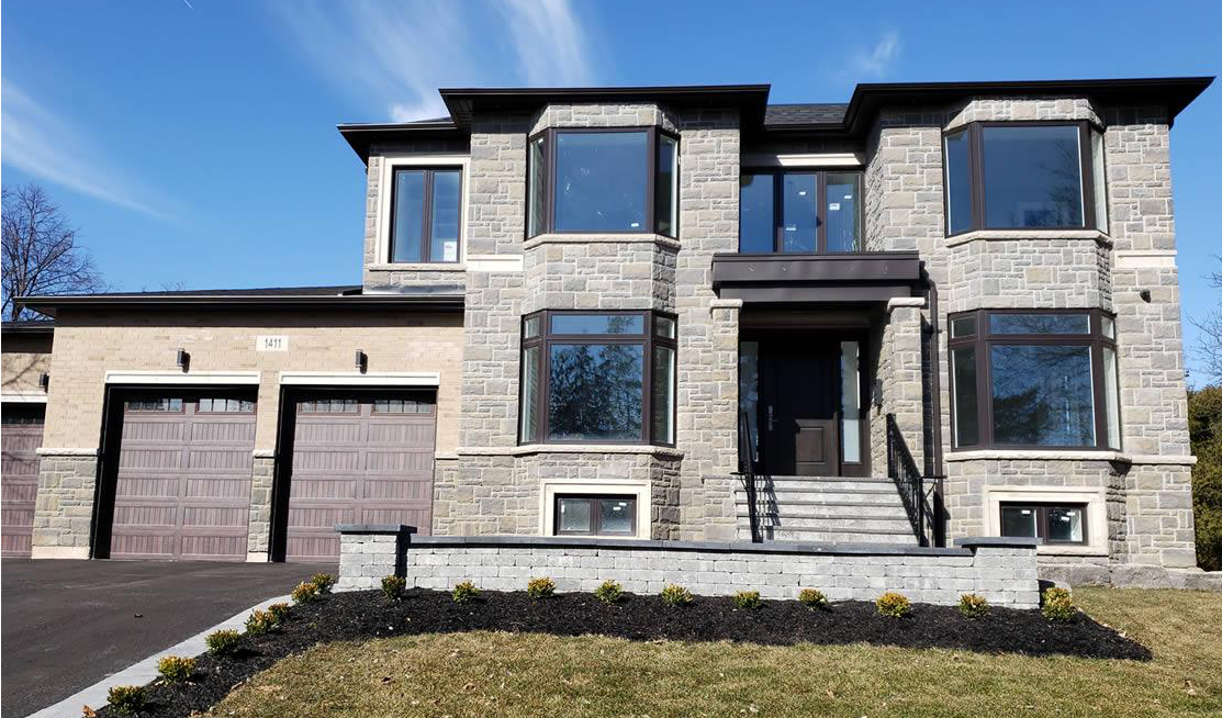 Two-story modern stone house with large bay windows, double garage doors, and a small landscaped front yard under a clear blue sky.