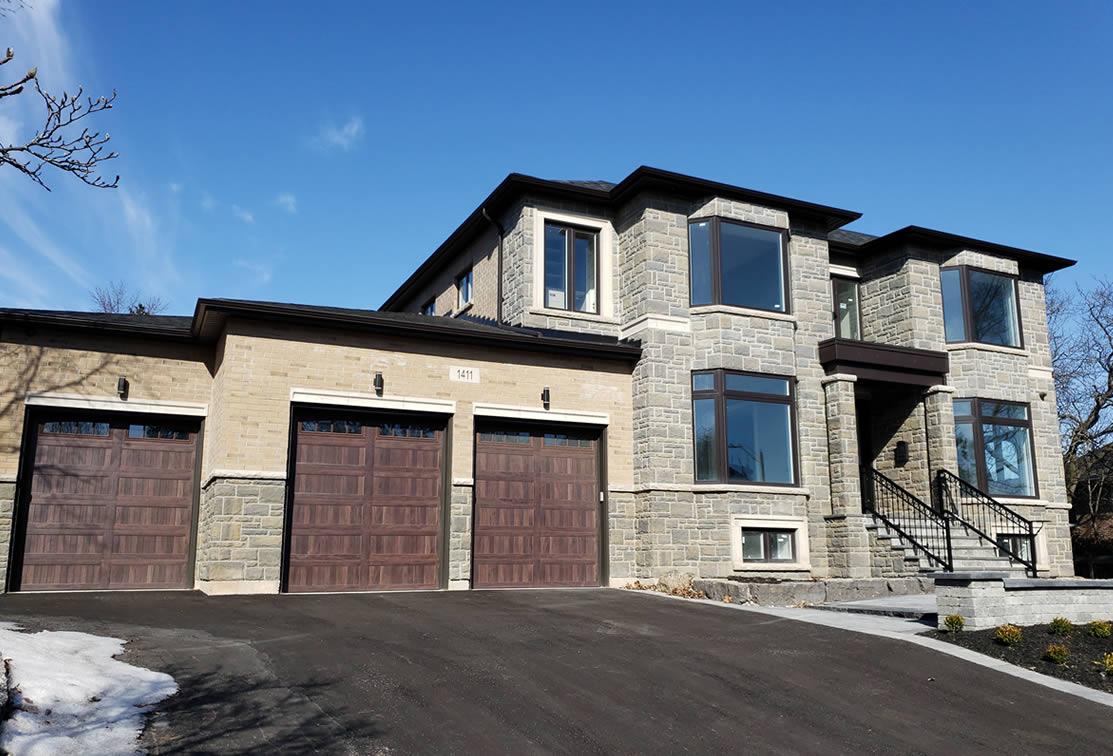 Two-story modern house with stone and brick exterior, large windows, and a three-car garage. Driveway and minimal landscaping are visible under a clear blue sky.