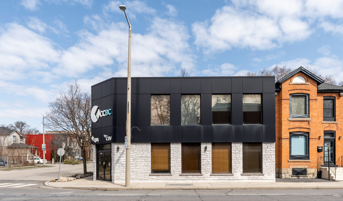 A modern two-story building with a black and white facade and large windows stands beside a red brick house on a quiet street at 374 Main Street West, under a partly cloudy sky.
