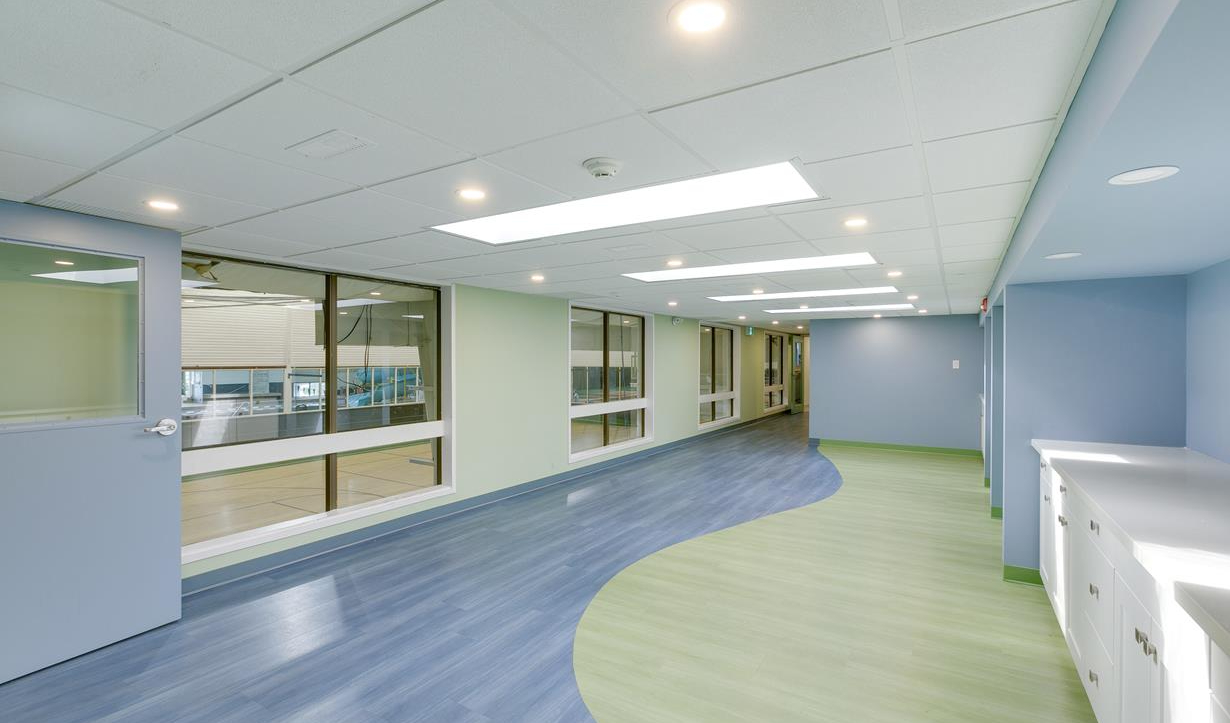 Bright hallway with blue and green flooring, white ceiling tiles, large windows on the left, and white cabinets on the right.