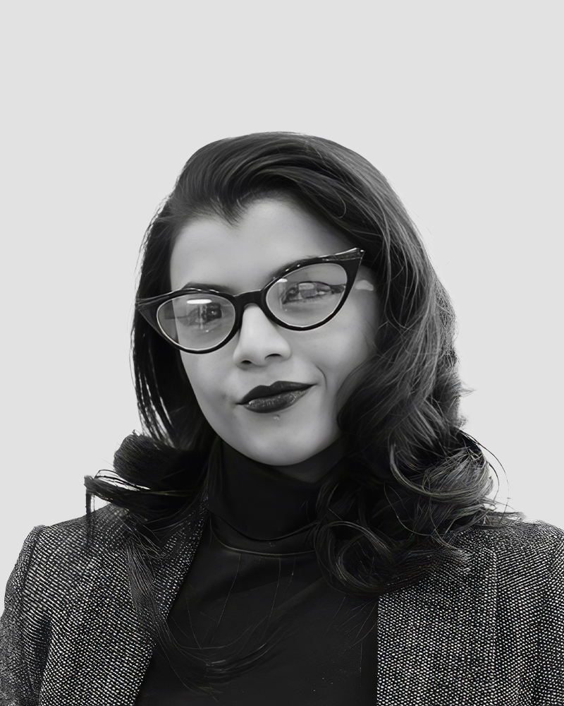 A person with dark hair wearing dark-rimmed glasses and a blazer.